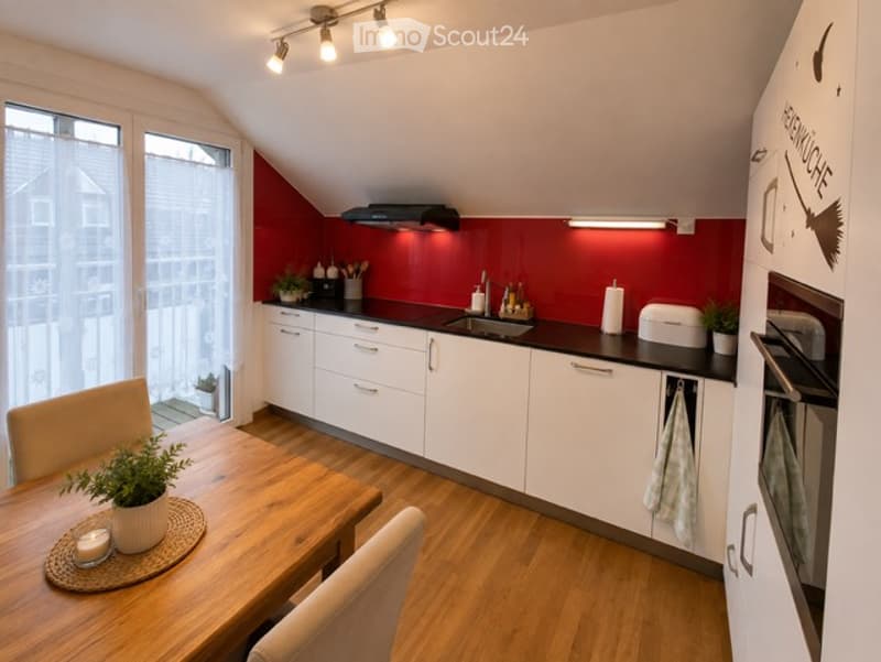 Modern kitchen with white cabinets, red accent wall, wooden floor, and a small dining area with a table and chairs. Features include a sink, oven, and a balcony view.