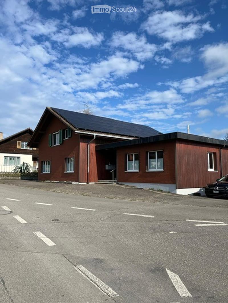 Two-story building with red brick facade, white trim, and a flat roof section. The building has multiple windows and a small entrance area.