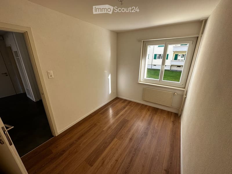 Empty room with wooden flooring, light-colored walls, a window with white frames, a radiator, and a door leading to another space.