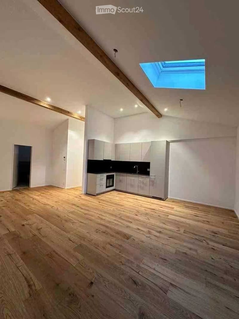 Empty room with wooden flooring, white walls, and a small kitchen area with cabinets and appliances.