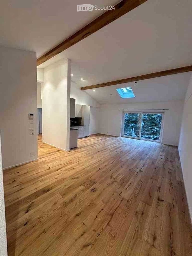 Empty room with wooden flooring, white walls, and large windows, featuring exposed wooden beams on the ceiling and a skylight.