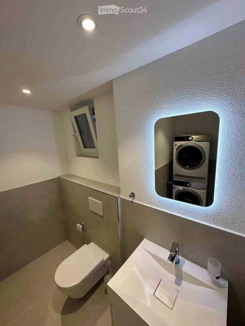 Modern bathroom with a toilet, sink, and a built-in washing machine. Features include a small window, a recessed light fixture, and a sleek, contemporary design.