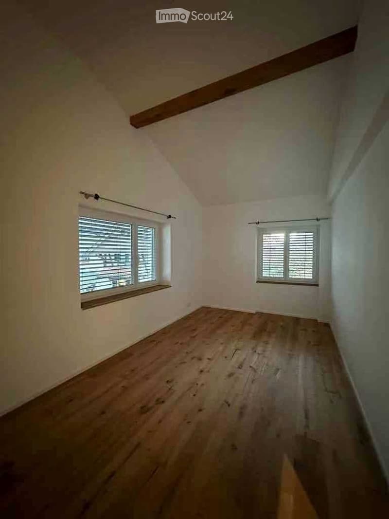 Empty room with wooden flooring, white walls, and two windows with blinds. Exposed wooden beams on the ceiling.