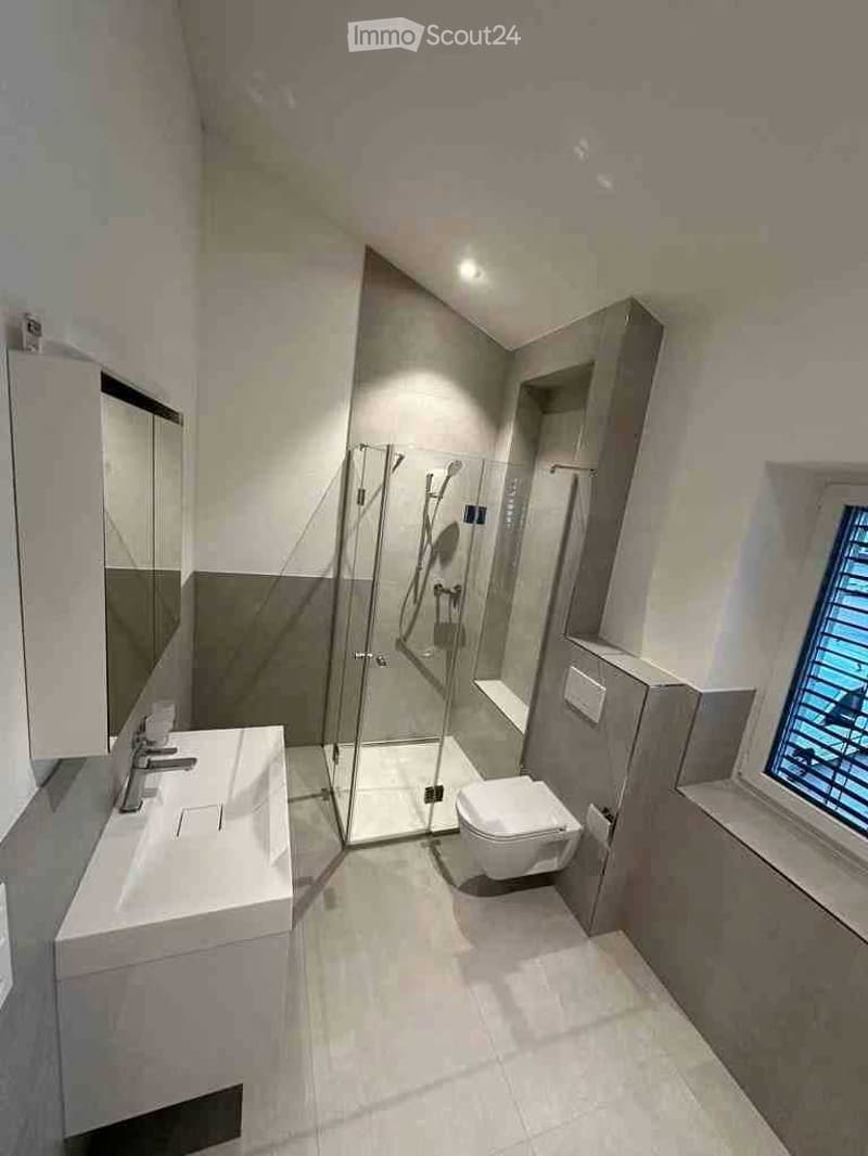 Modern bathroom with white tiles, a glass-enclosed shower, a toilet, a sink with a mirror, and a window with blinds.