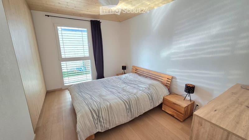 Bedroom with wooden floor, white walls, wooden bed, wooden nightstands, lamps, large window with blinds, wooden ceiling