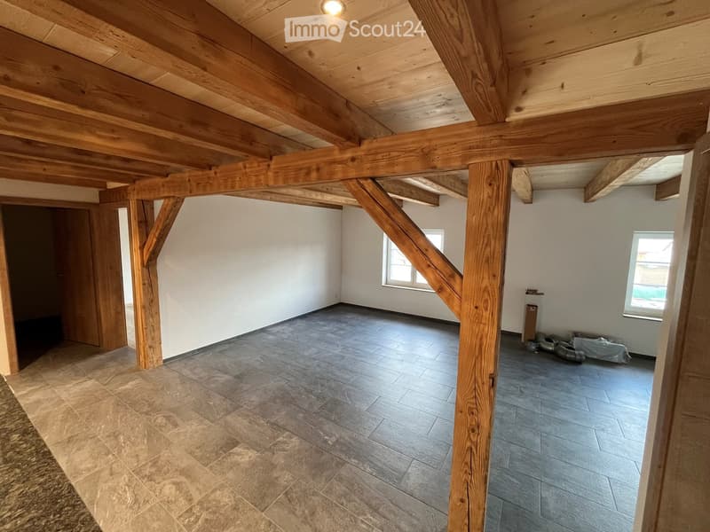 Empty room with exposed wooden beams, tiled floor, and white walls. Two windows and a door are visible.