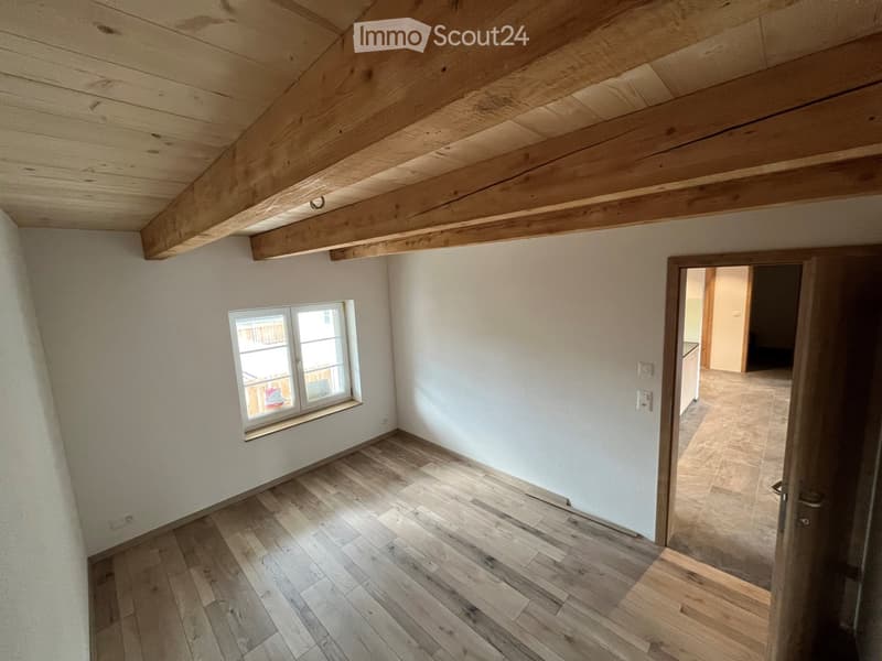Empty room with wooden floor, white walls, exposed wooden ceiling beams, and a small window with white frame. A doorway leads to another room with tiled flooring.