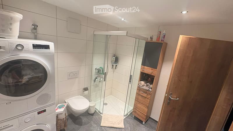 Modern bathroom with a toilet, glass-enclosed shower, Bosch washing machine, wooden cabinet, and various toiletries.