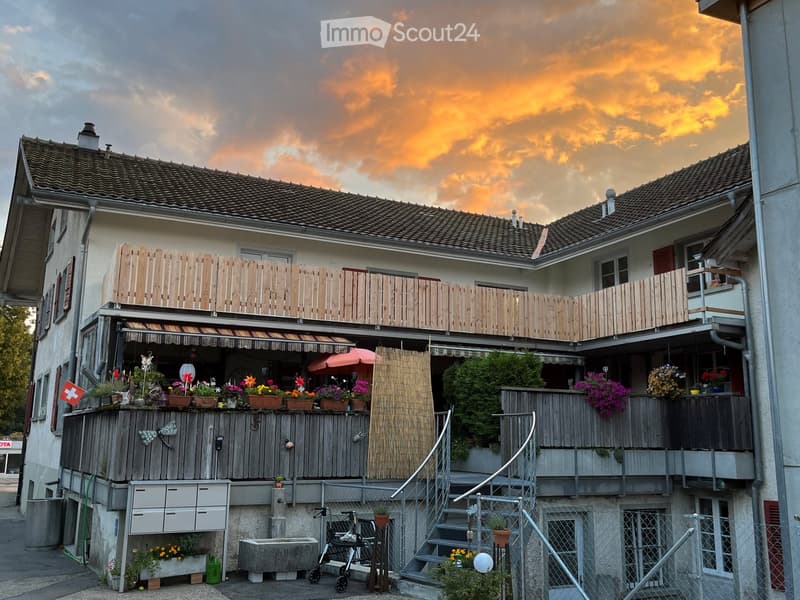 A multi-story residential building with balconies, wooden railings, and flower pots, set against a sunset sky.