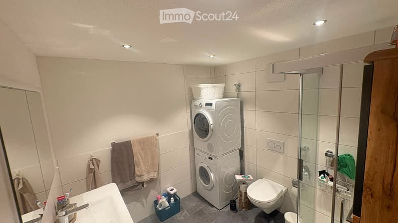 Modern bathroom with a white tiled shower, a toilet, a sink, and a stacked washing machine and dryer. Towels are hanging on a rack, and a small basket holds cleaning supplies.