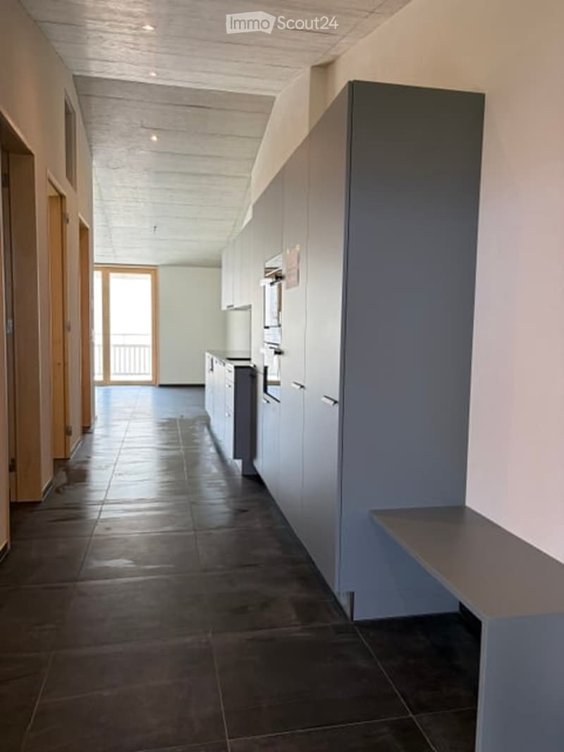 Modern kitchen area with fitted cabinets, dark tiled floor, and a small bench near the cabinets. The space is bright with natural light from a large window and a balcony door.