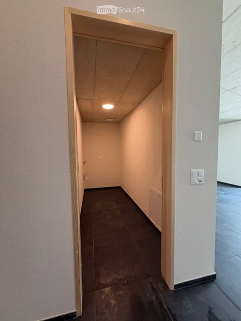 Empty hallway with beige walls, dark tiled floor, and a single ceiling light. The space is clean and unfurnished, with no visible furniture or decorations.