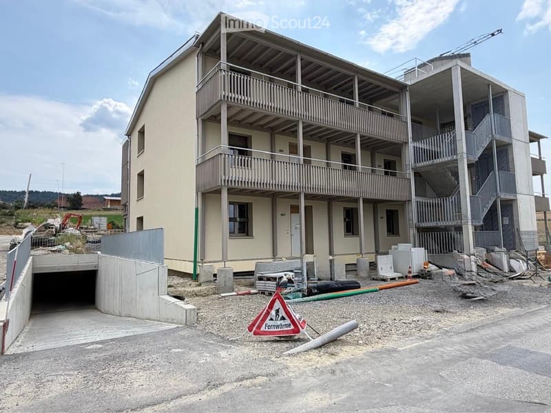 Multi-story residential building under construction with exposed balconies, a covered parking area, and construction materials scattered around.