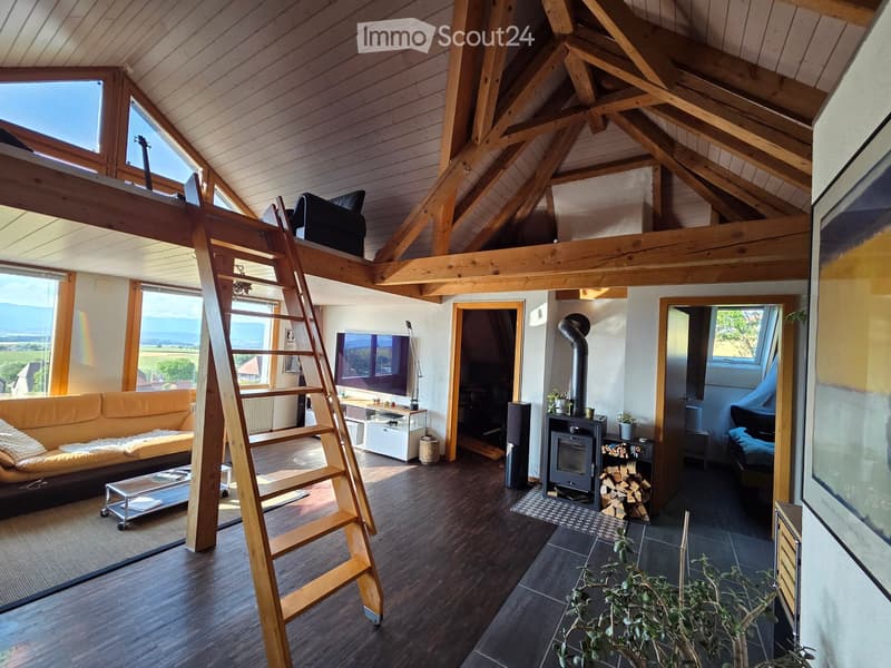Bright living room with wooden beams, exposed ceiling, large windows offering a scenic view, a fireplace, a sofa, coffee table, television, and a small loft area accessible by a wooden ladder.