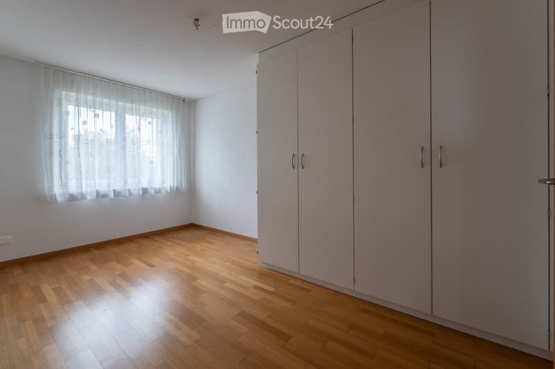 Empty room with wooden flooring, white walls, large window with sheer curtains, and built-in closet with double doors.