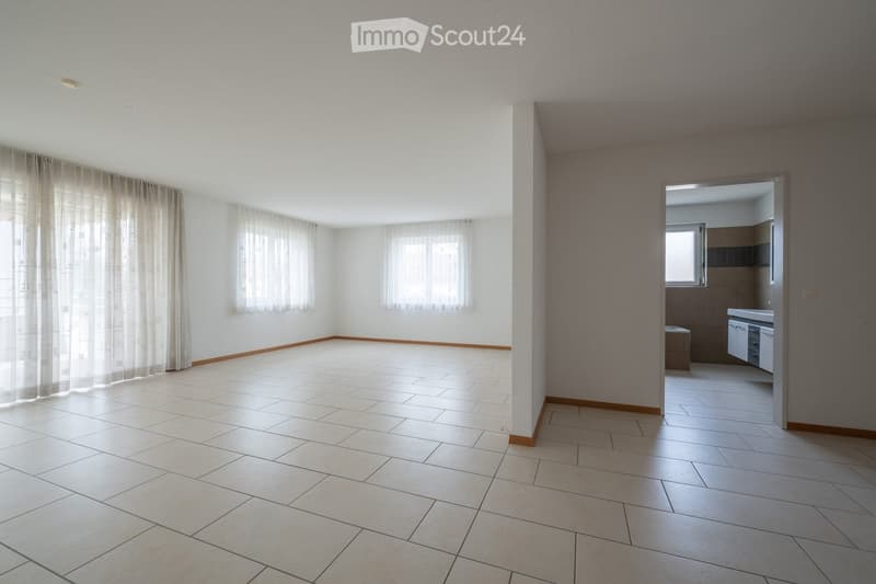 Empty room with tiled floor, white walls, large windows with sheer curtains, and a doorway leading to a small kitchen area.