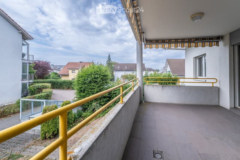 Balcony with tiled floor, yellow railings, and striped awning, overlooking a residential area with houses and greenery.