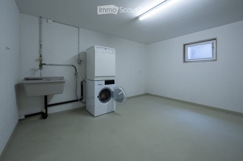 A small utility room with a washing machine, dishwasher, and a sink, featuring white tiled walls and a concrete floor.