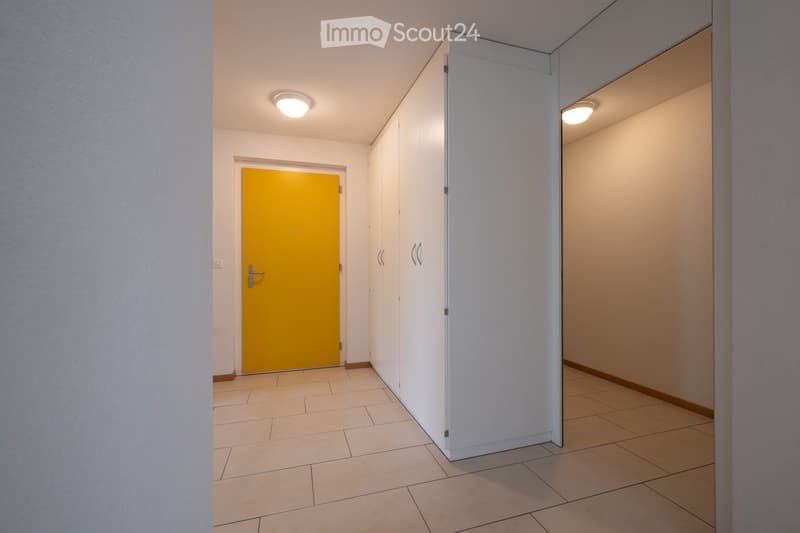 A clean, modern hallway with tiled flooring, white walls, and a bright yellow door at one end. Recessed ceiling lights provide illumination, and built-in white cabinets line one wall.