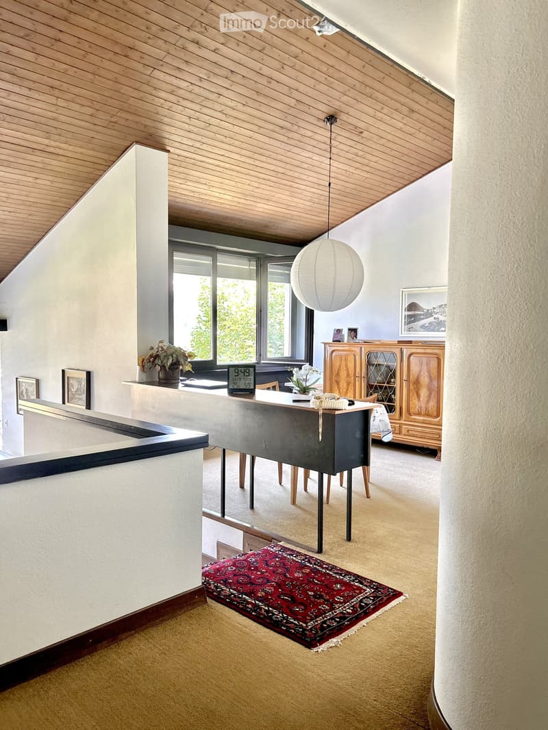 Modern living room with a slanted wooden ceiling, large windows, a round ball lamp, a wooden cabinet, a table with chairs, and a patterned rug.
