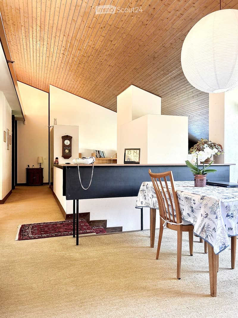 A cozy, well-lit room with a slanted wooden ceiling, a dining table with a floral tablecloth, a wooden chair, a clock on the wall, a laptop on the table, a flower vase, a small cabinet, and a lamp on a side table.