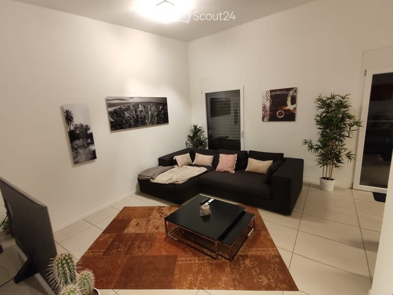 Modern living room with a black sectional sofa, coffee table, cushions, and decorative items. Features a large window with blinds, artwork on the walls, and potted plants.