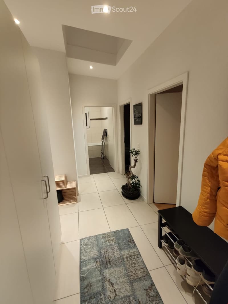 A clean, modern hallway with white tiled flooring, white walls, and several doors leading to different rooms. A small plant sits near the center, and a shoe rack with multiple pairs of shoes is visible on the right side.