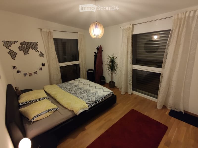 Cozy bedroom with a double bed, yellow pillows, patterned duvet, wooden floor, large window with curtains, and a hanging lamp.