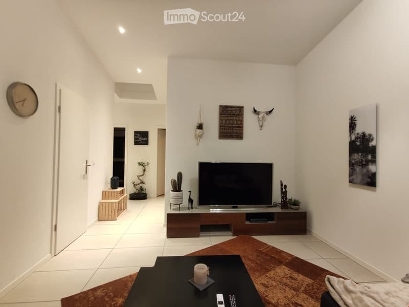 Modern living room with white walls, tiled floor, television on a wooden stand, coffee table with remote controls, decorative items including a clock, picture frames, and plants.