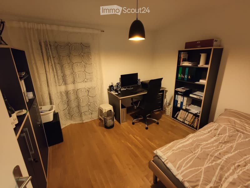 Cozy bedroom with a bed, desk, bookshelf, and a hanging lamp. The room features wooden flooring and a patterned curtain.