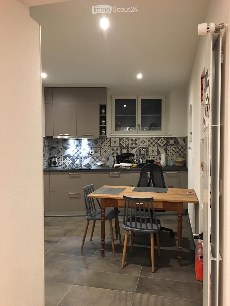 Modern kitchen with grey cabinets, a wooden dining table with four chairs, a sink with a faucet, and various appliances including a blender and a refrigerator.