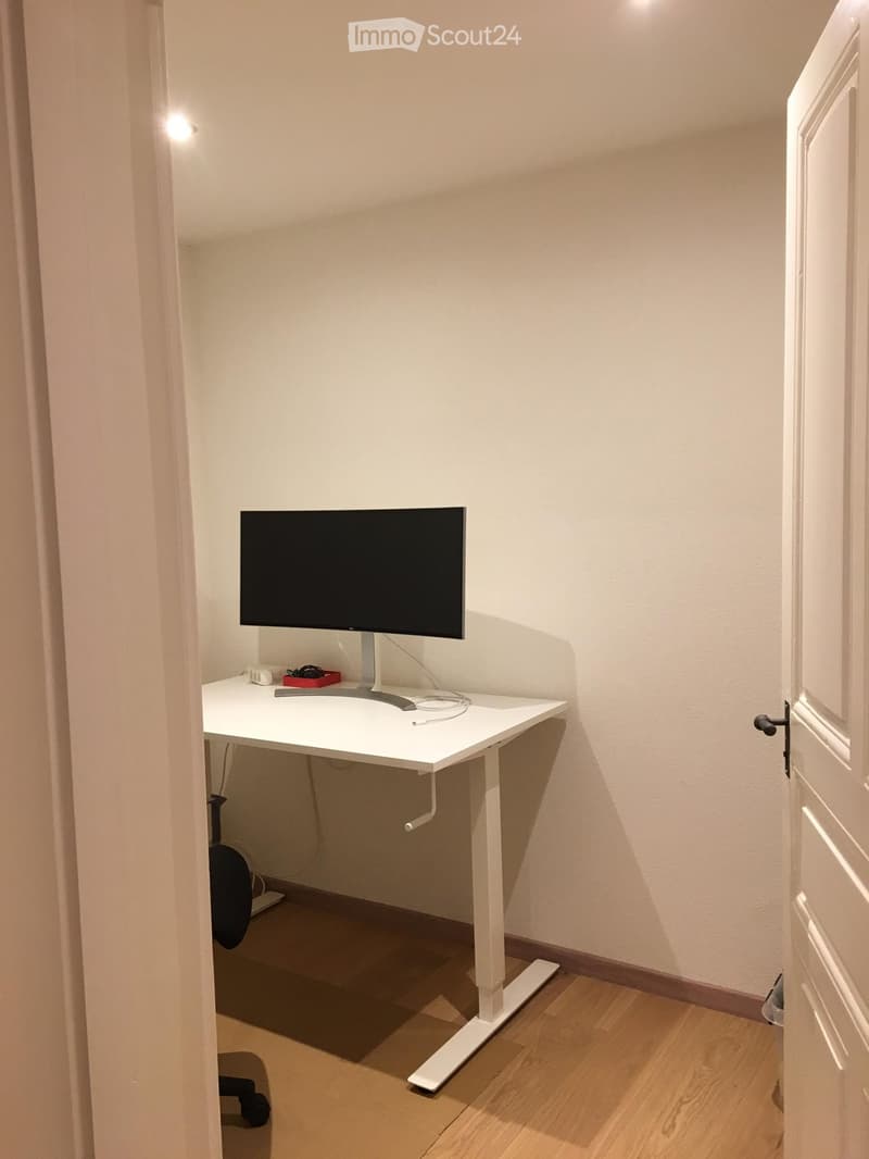 A small, well-lit room with a white desk, a black monitor, and a black chair tucked under the desk.