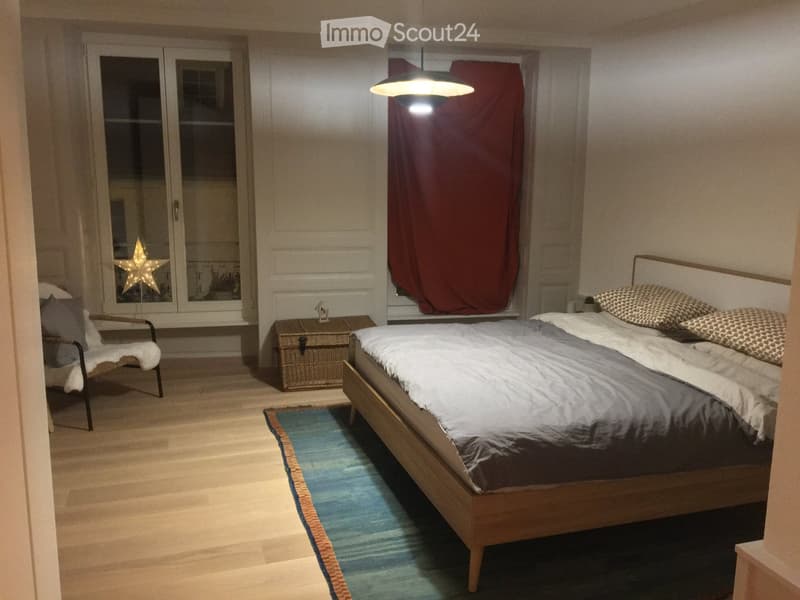 Cozy bedroom with a wooden bed, patterned pillows, a wicker trunk, a blue rug, and a red curtain on the window.