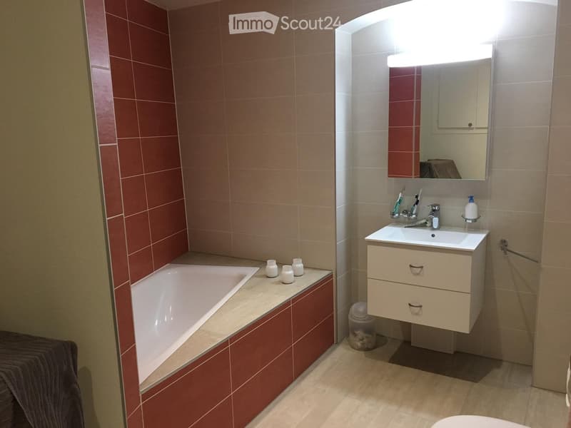 Modern bathroom with red and beige tiles, a bathtub, a sink with a cabinet, a mirror with built-in lighting, and a towel on the edge of the bathtub.