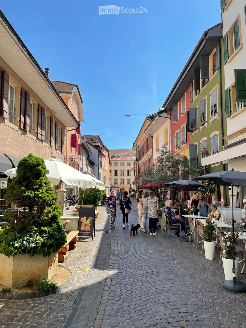 A charming cobblestone street lined with colorful buildings, outdoor seating areas, and people walking. The street is adorned with potted plants, umbrellas, and bicycles.