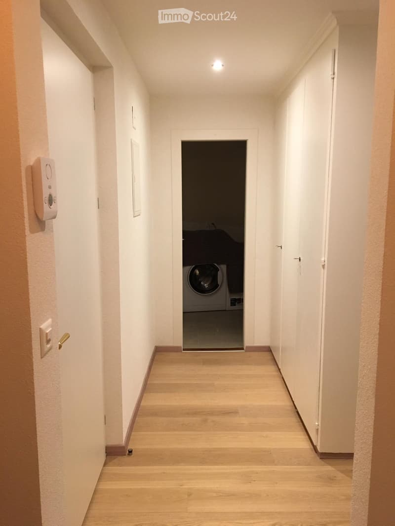 A narrow hallway with light wooden flooring, white walls, and a recessed ceiling light. An open doorway leads to a laundry area with a washing machine visible.
