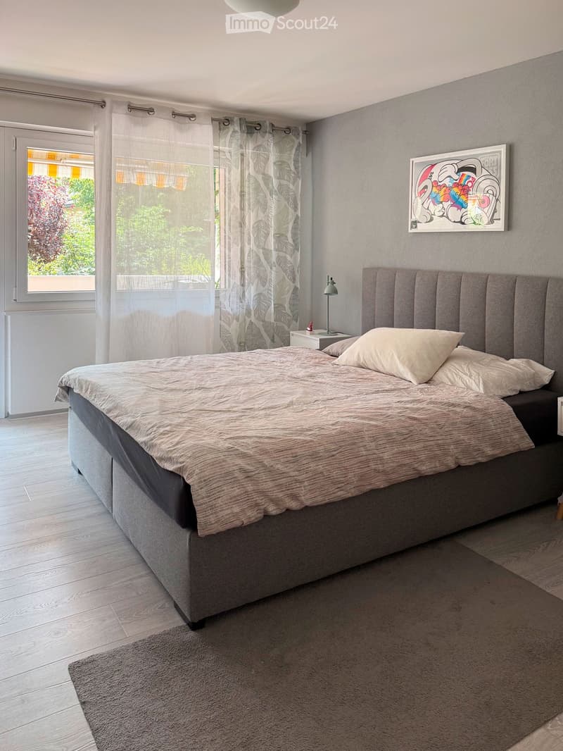 Modern bedroom with a large bed, nightstand, lamp, and a colorful picture frame on the wall. The room features light wooden flooring and sheer curtains on the window.