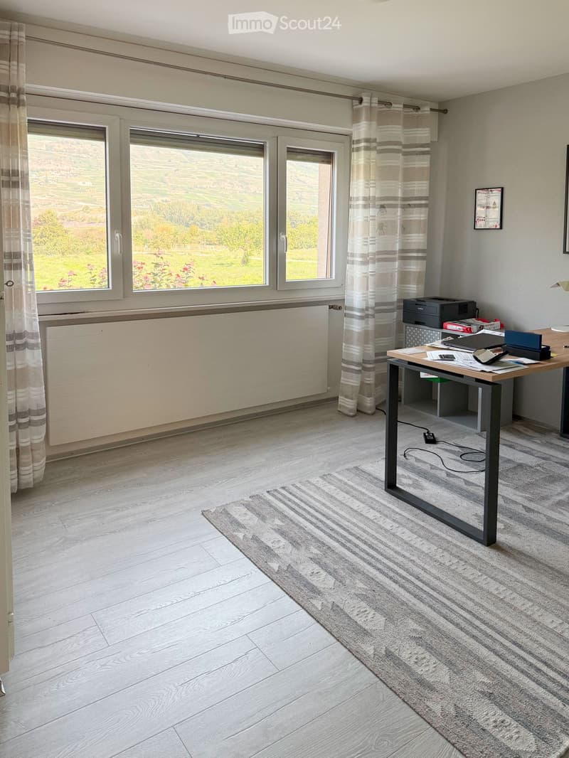 Bright room with wooden flooring, large windows with curtains, a desk with a laptop, printer, and other office supplies, and a small rug.