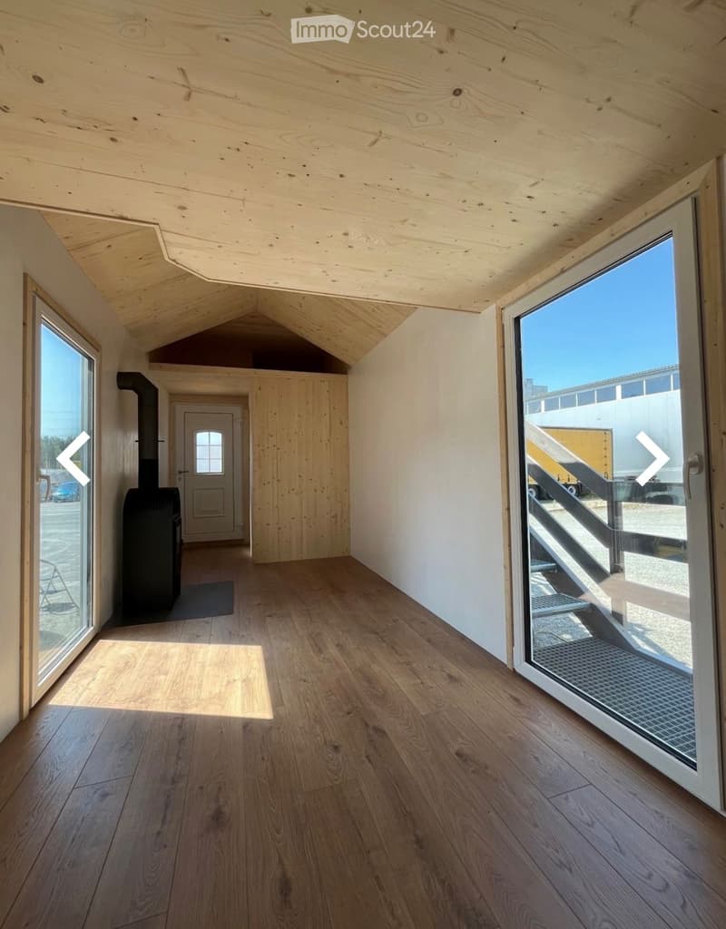 Empty room with wooden flooring, white walls, and large windows. Features a fireplace and a door leading to another room.