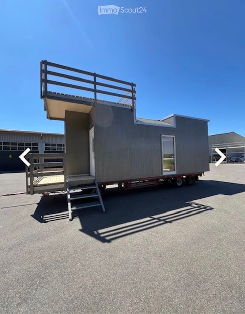 A modern, grey, prefabricated tiny home on wheels, featuring a raised deck with a wooden railing and a staircase leading up to the entrance.