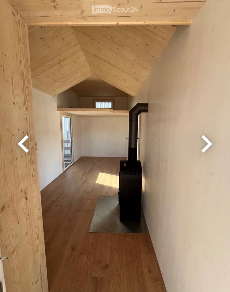 A narrow hallway with wooden flooring, white walls, and a black wood-burning stove in the center. The ceiling has exposed wooden beams and a slanted design, with a small window at the far end.