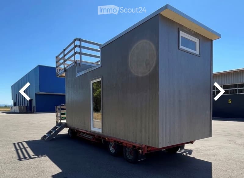 A modern, compact, grey wooden cabin on wheels with a raised deck and large windows, parked on a concrete surface.