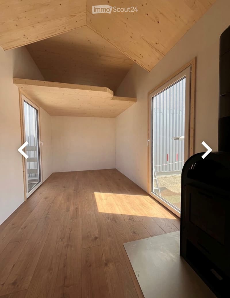 Empty room with wooden flooring and white walls, featuring large windows and a slanted wooden ceiling.