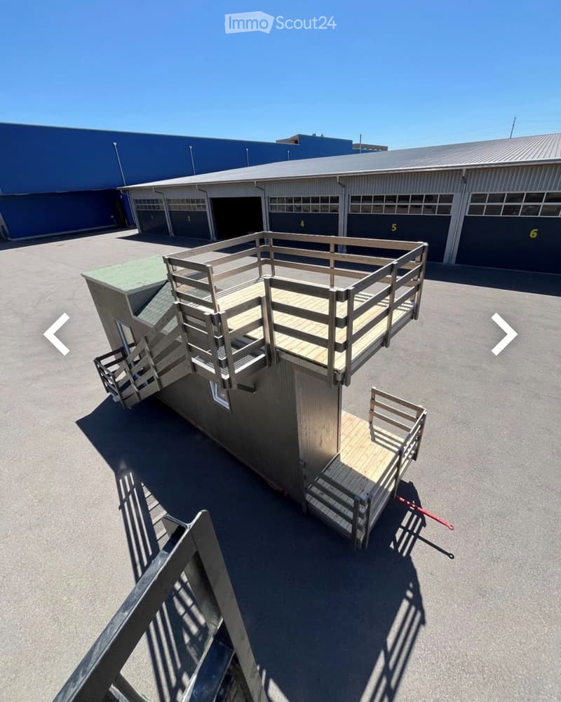 A small, modern wooden structure with a raised platform and stairs, situated in an industrial area with large warehouses in the background.