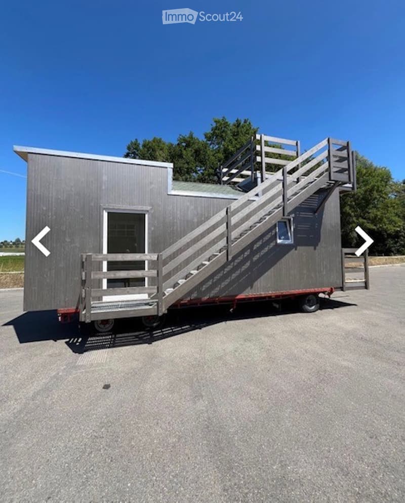 A small, modern, grey trailer-style building with a staircase leading to an upper level, parked on a paved surface.