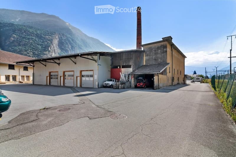 Industrial building with large garage doors, a tall chimney, and a mountainous backdrop. Vehicles are parked in front, and the area includes a paved driveway.