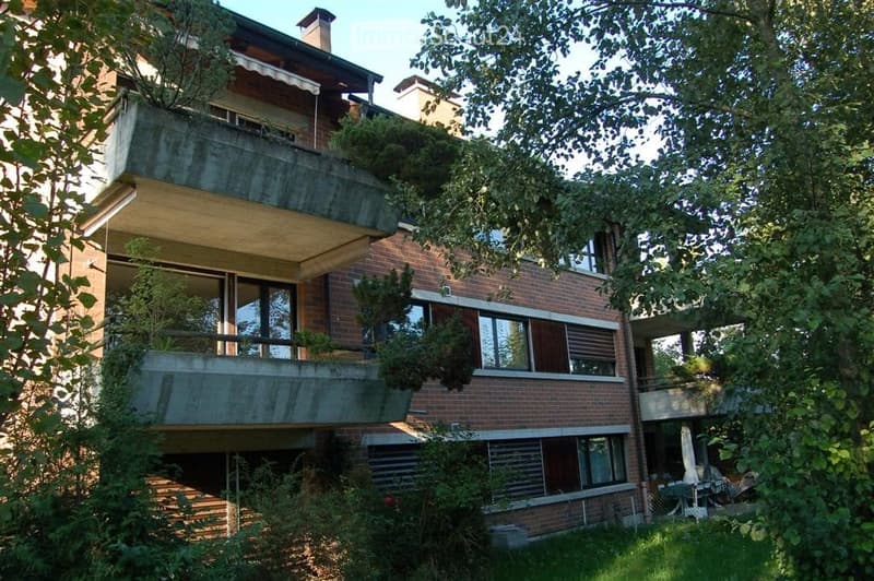 Multi-story brick building with large windows and balconies, surrounded by greenery and trees.