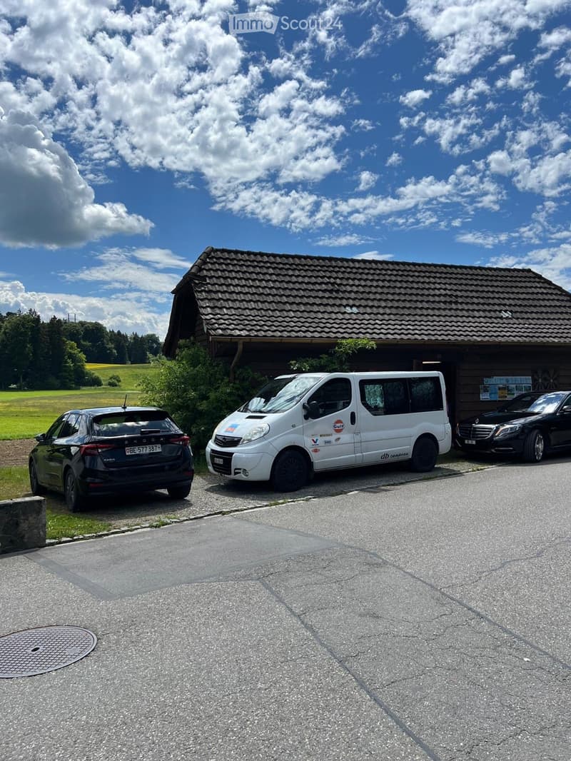 A small wooden building with a tiled roof, surrounded by a grassy field and trees. Three vehicles are parked in front of the building on a paved area.