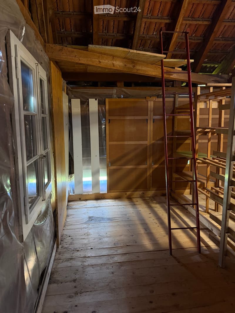 Empty attic space with wooden floors and exposed wooden beams, featuring a window on the left and a ladder leading to the upper level.