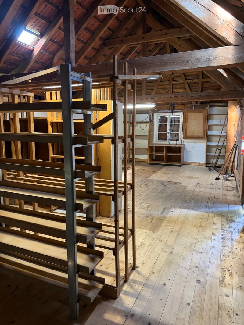 An attic space with exposed wooden beams and rafters, wooden floor, and a small window. The room is empty with no furniture or furnishings.